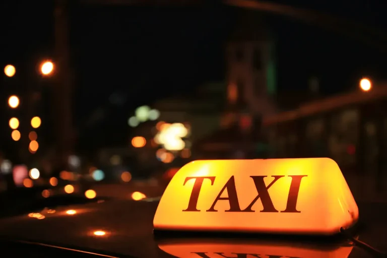 Lit taxi sign on a car roof at night with city lights behind it