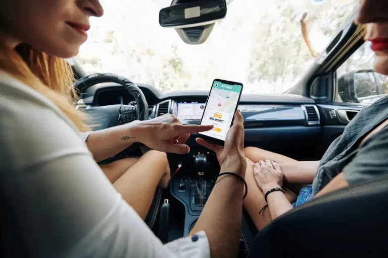 Car passenger holding a phone with a ride app open while the driver waits at the wheel